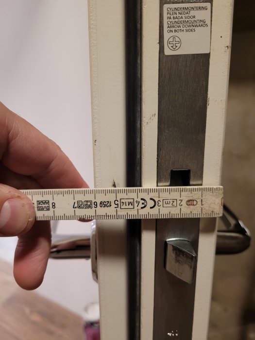 Hand med tumstock mäter dörrlåsets bredd vid instruktionsklistermärke om cylinderinstallation.