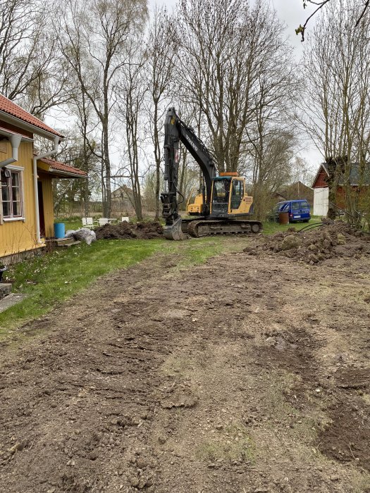 Grävmaskin på ett uppgrävt landområde, nära hus och träd, grå molnig himmel, orörd natur i bakgrunden.