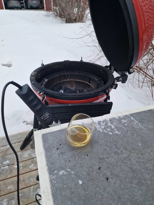 Röd keramisk grill med lock öppet, aska inne, uteplats, snötäckt trädgård, dricksglas på bordet.