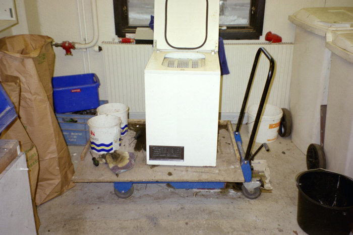 Tvättmaskin på en blå pallvagn i rörigt, underhållsutrymme med byggmaterial och högtryckstvätt.