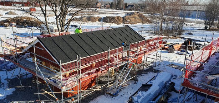 Byggarbetsplats med rött hus, takläggning, snö, byggställningar och arbetare i säkerhetsväst.