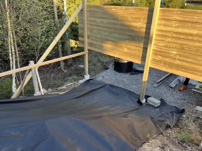 Trädäck under konstruktion med stödstag, svart markduk, grus, och bakgrund av skog.