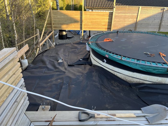Utomhus byggarbetsplats, trampolin, konstruktionstimmer, byggmaterial, verktyg, svart markduk, staket, skymning.