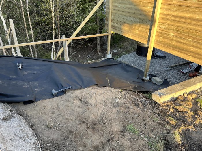 Byggplats med svart markduk, träkonstruktion, verktyg och natur omkring i dagsljus.