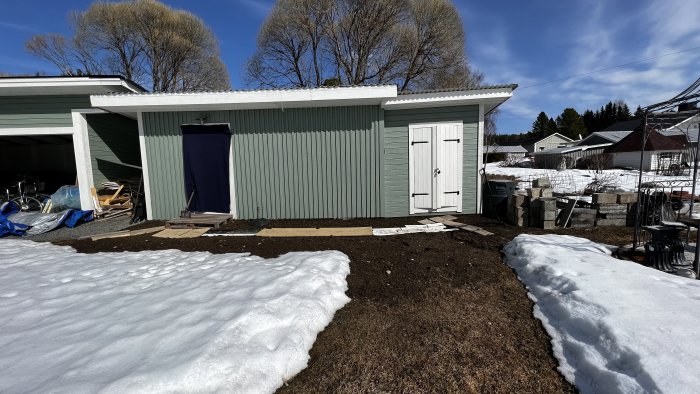 Bakgård med carport och skjul, smältande snö, blå himmel, tidig vår, oordnade föremål, grönsakande träd.