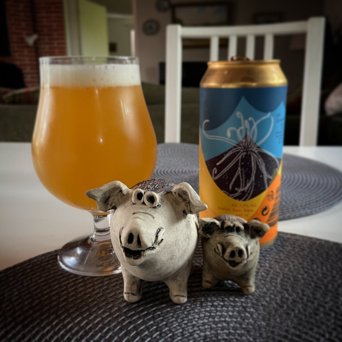 Ölglas med grumlig vätska, ölburk, två små griskulpturer på bord med diskret bakgrund.