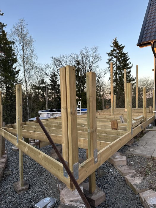 Under uppbyggnad träveranda, stödpelare, bjälklag, blå himmel, träd, grus, betongplattor.