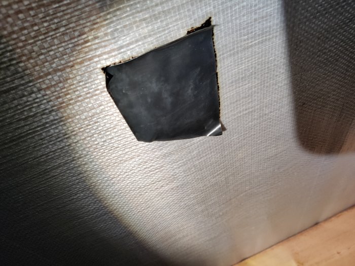 Hål i vävd vägg, synlig mörk insida, skadad textil, slitet, närbild, ljusskiftningar, ojämn kant.