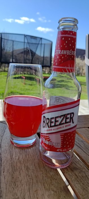 Flask Breezer Jordgubb, glas med röd dryck, träbord, utomhusmiljö med grönt gräs, soligt, avslappnat.