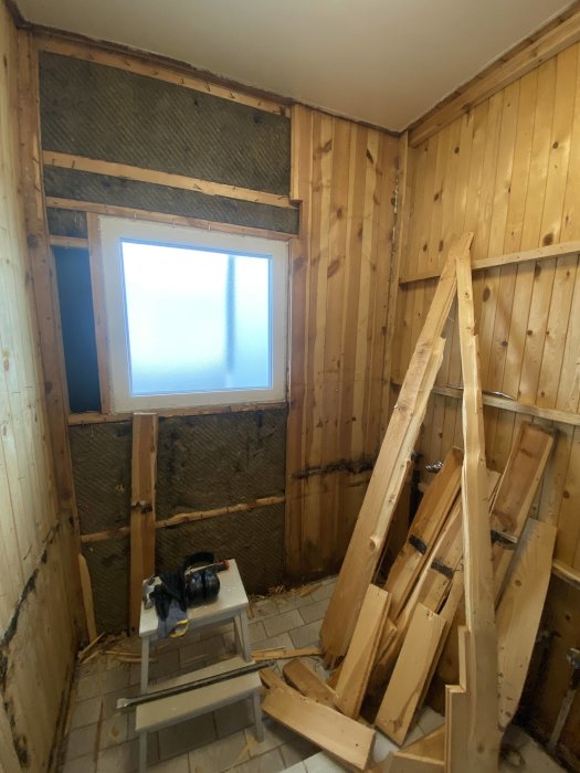 Rum under renovering, isolering synlig, träpanel, rörigt, byggmaterial och verktyg på golvet.
