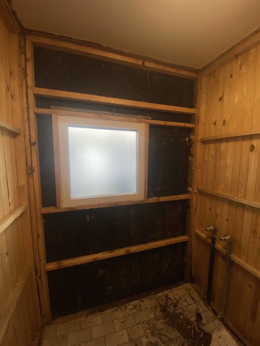Tomt rum med träpaneler, frostat fönster, renovering behövs, mörkt, skräpigt golv, avskalad vägg.