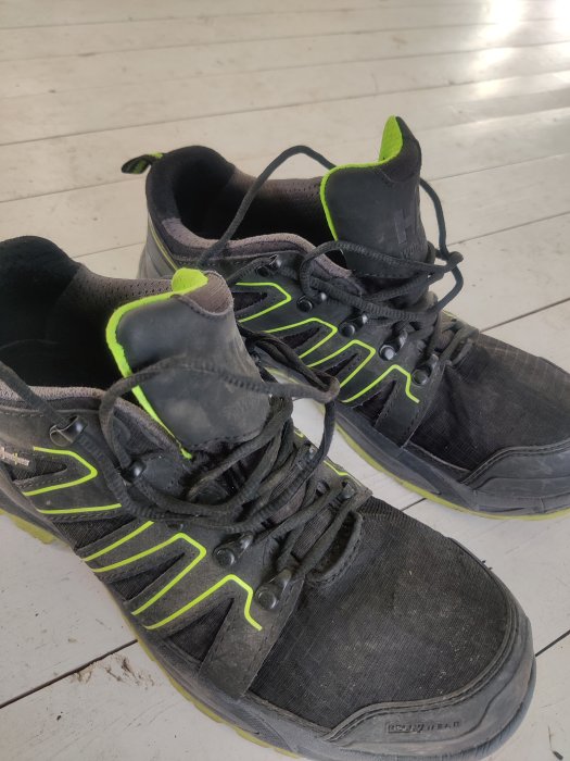 Ett par slitna svarta sneakers med neon-gröna detaljer på ett trägolv.