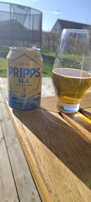 Ölburk och glas på träbord utomhus i soligt väder, med grön bakgrund, Pripps Blå Citrus, avslappnad atmosfär.
