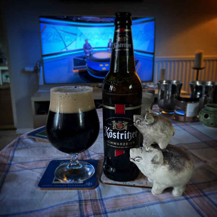 Ölflaska, ölglas med mörkt öl, grisskulpturer, TV i bakgrunden med suddig bild.