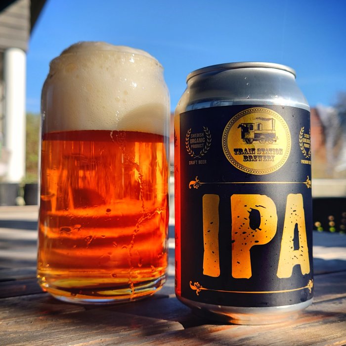 Ett glas öl med skum bredvid en IPA-ölburk utomhus, solsken, träyta.