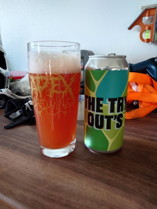 Ett glas med skummande röd-orange dryck och en färgglad burk med text “THE TRY OUT’S” på ett bord.