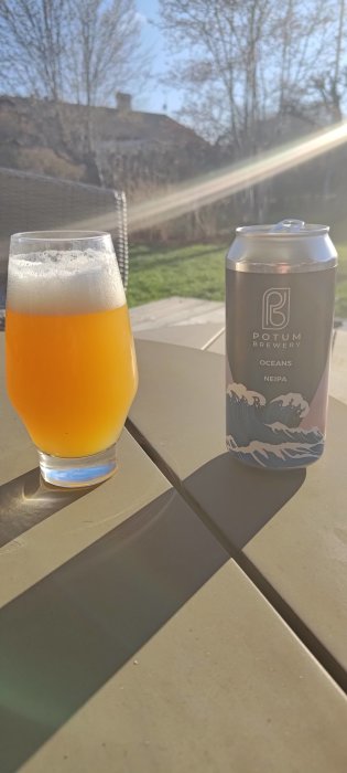 Ölglas med skum på utebord, burk bredvid, soligt, träd och blå himmel bakom.