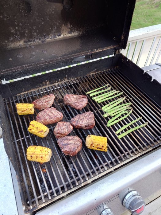 Gasolgrill med grillade biffar, majs och sparris utomhus på en balkong.