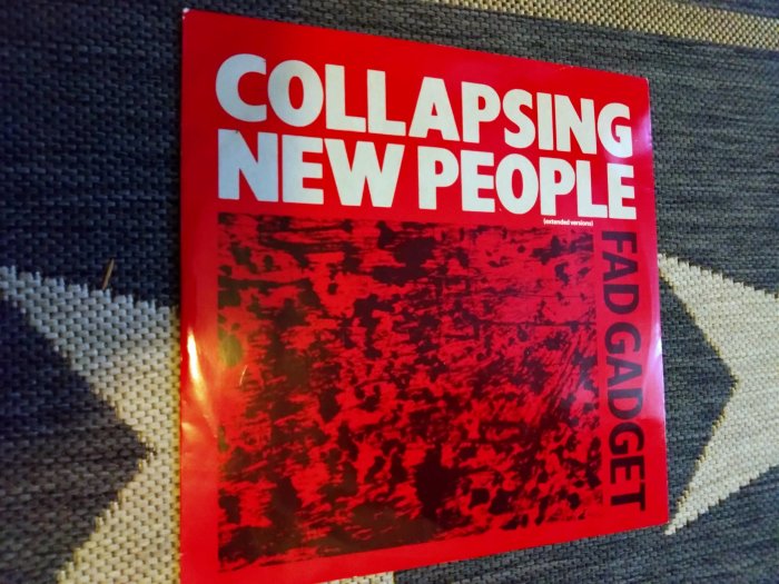 Röd skivomslag för "Collapsing New People" av Fad Gadget, extended versions, ligger på tyg.