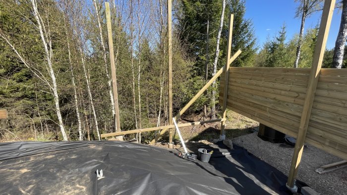 Byggarbetsplats med trästomme, verktyg, lövträd, soligt väder, svart markduk, ute i naturen.