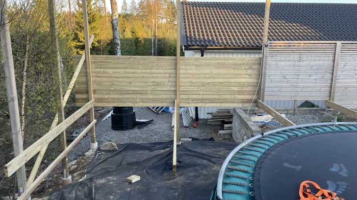 Trädgård under konstruktion med staket, byggmaterial, trampolin och delar av hus.
