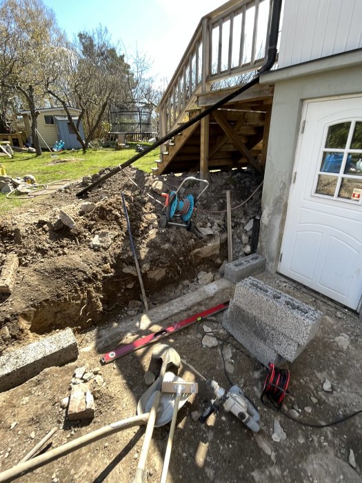 Trädgård med grävd grop, trätrapp, verktyg, och byggmaterial vid husgrund under renovering eller konstruktion.