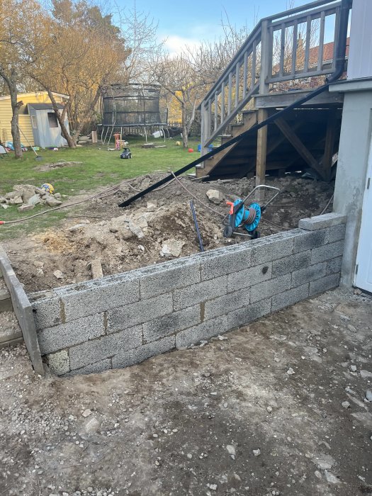 Grävd trädgård, murblocksvägg under konstruktion, trädäck, leksaker i bakgrunden, slangvinda, grått grus.