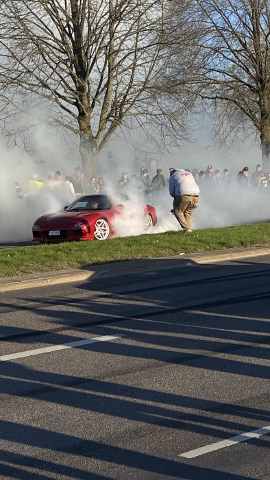 Röd bil gör burnout, tjock rök, åskådare i bakgrunden, soligt väder, träd utan löv.