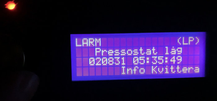 Digital display med text "Larm Pressostat låg" och en tidsstämpel, i mörker med rött ljus ovanför.