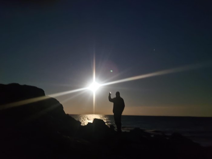 En silhuett av en person på klippor vid havet med lysande ljusstråle i skymningen.