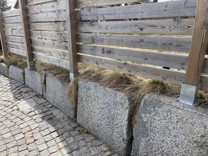 Trästaket monterat på granitblock med gräs, vid kullerstensbelagd gata. Soligt väder.