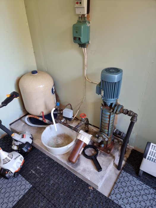 Vattenpump och filter i ett hörn, arbetsverktyg och utrustning, elektrisk grön mätare på väggen.