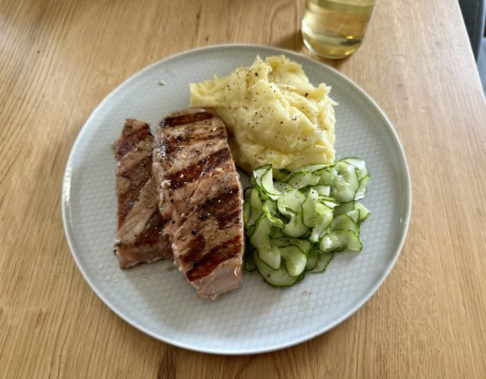 Grillad köttbit, mosad potatis, gurksallad på tallrik, bord, dryck i glas.