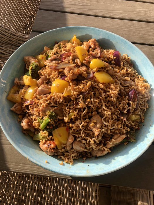 Stir-fried noodles with chicken, broccoli, bell peppers, and onions in a bowl, outdoors on a sunny day.