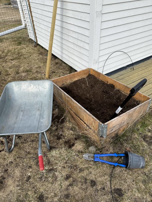 Trädgårdsbild: kärra, odlingslåda med jord, spade, räfsa och grensax. Trädgårdsarbete eller förberedelse för plantering.