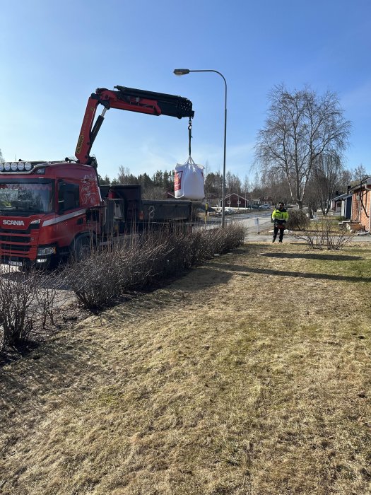 Röd lastbil med kran lyfter stor vit säck, person i säkerhetskläder observerar, soligt, bostadsområde i bakgrunden.