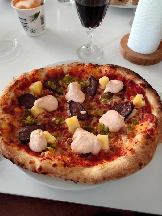 En pizza med ananas, kött, grönsaker, rosa sås, på en vit tallrik, rödvin i glas, bakgrund av köksinteriör.