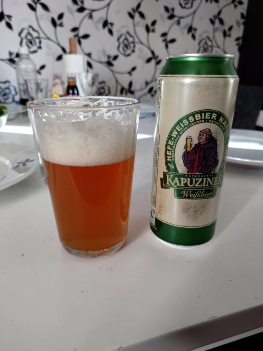 Ett glas med skummande öl och en burk Kapuziner Weißbier på ett bord.