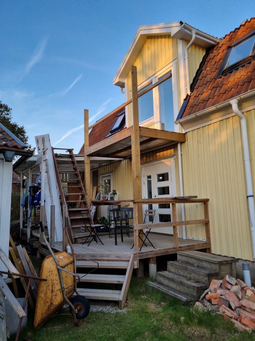 Gult hus med veranda under konstruktion, trädgårdsredskap, kvällssol, öppen dörr, klädstreck, byggmaterial.