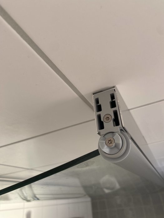 Glashylla i hörn med metallbeslag, vita kakelväggar, skruvförband och skenor syns.