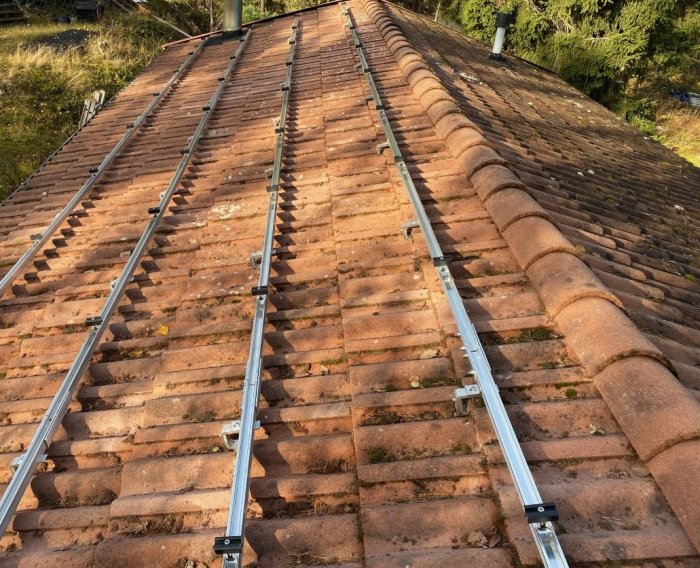 Tegelrött tak med monterade metallräls, möjligtvis för solpaneler, mot solig skogsbakgrund.