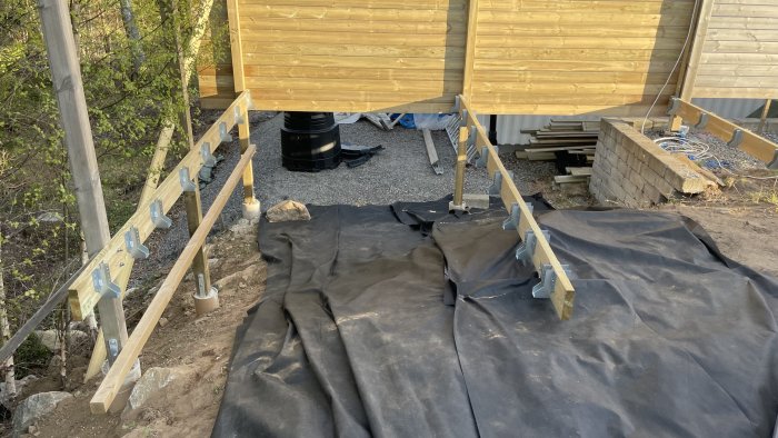 Byggarbetsplats med stödben, svart markduk, konstruktionsmaterial och verktyg; oavslutat byggprojekt utomhus.