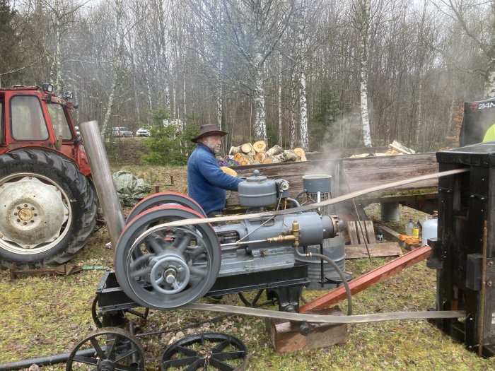 Man med hatt använder stationär ångmaskin nära röd traktor i skogsmiljö; träbearbetning med historisk teknik.