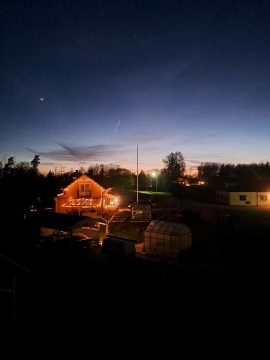 Kvällshimmel över bostadsområde med upplysta hus, växthus i förgrunden, gradvis solnedgång i bakgrunden.