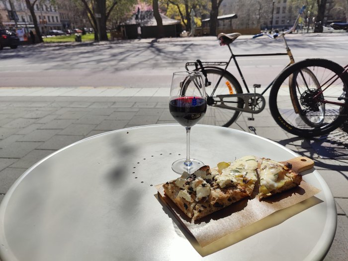Vin i glas, bröd med pålägg, uteservering, cykel i bakgrunden, soligt, stadsmiljö.
