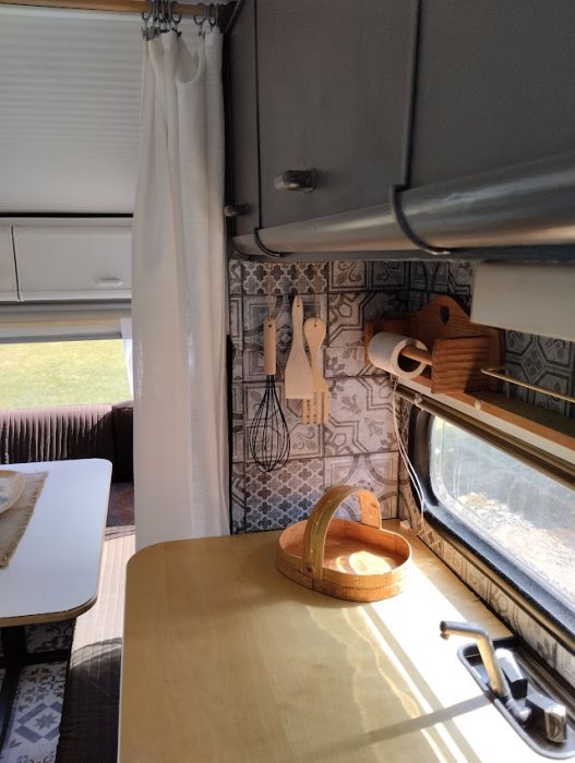 Husvagnsinteriör med köksredskap, bord, bänk och fönster, ljusinläpp, mysig, kompakt boendemiljö.