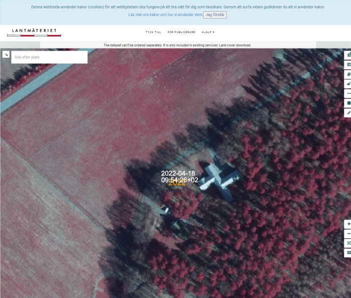 Flygbild över landskap med rödtonade fält och skog, byggnad, webbsidesinterface från Lantmäteriet. Datumstämpel synlig.