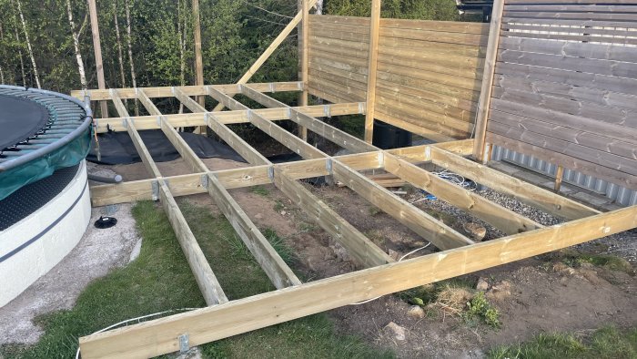 Trallkonstruktion under uppbyggnad, träreglar monterade, terränganpassning, del av studsmatta, staket, utomhusprojekt.
