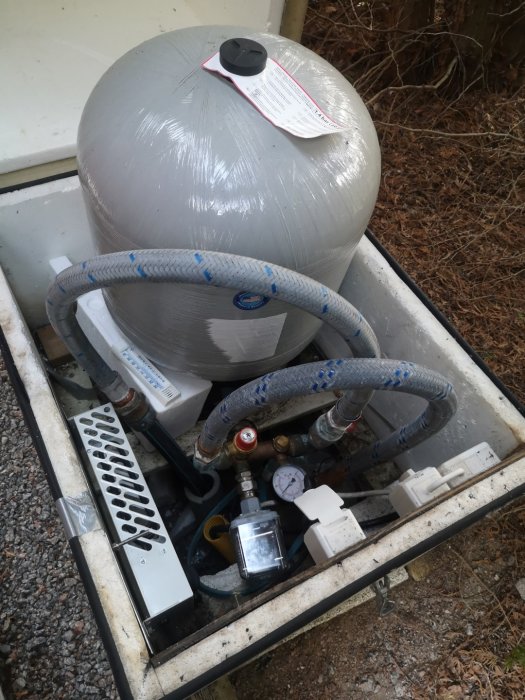 Poolfilter och pumpsystem med slangar, mätare och ventiler, uppställt på grusig mark.
