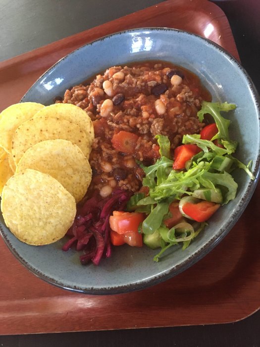 Chili con carne med bönor, tacochips, sallad, rödlök, servers på blå-grå tallrik. Vibrant, inbjudande, hemlagad måltid.